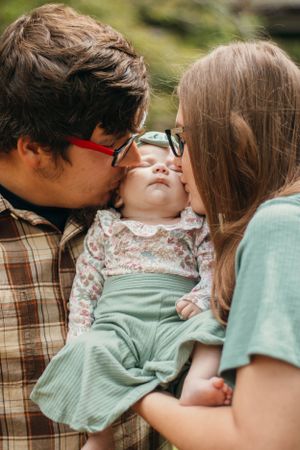 Parents giving gentle kisses to baby in floral outfit outdoors.