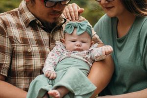 Baby in mint and floral outfit being held by adults in green and plaid clothing.