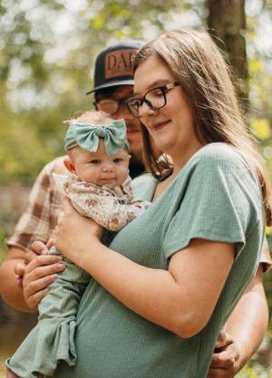 A couple in a green shirt and plaid holds their baby wearing a mint bow in an outdoor setting.