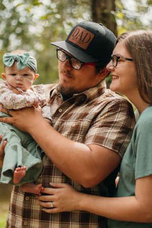 Happy couple wearing glasses and casual attire snuggle with their infant in a forest setting.