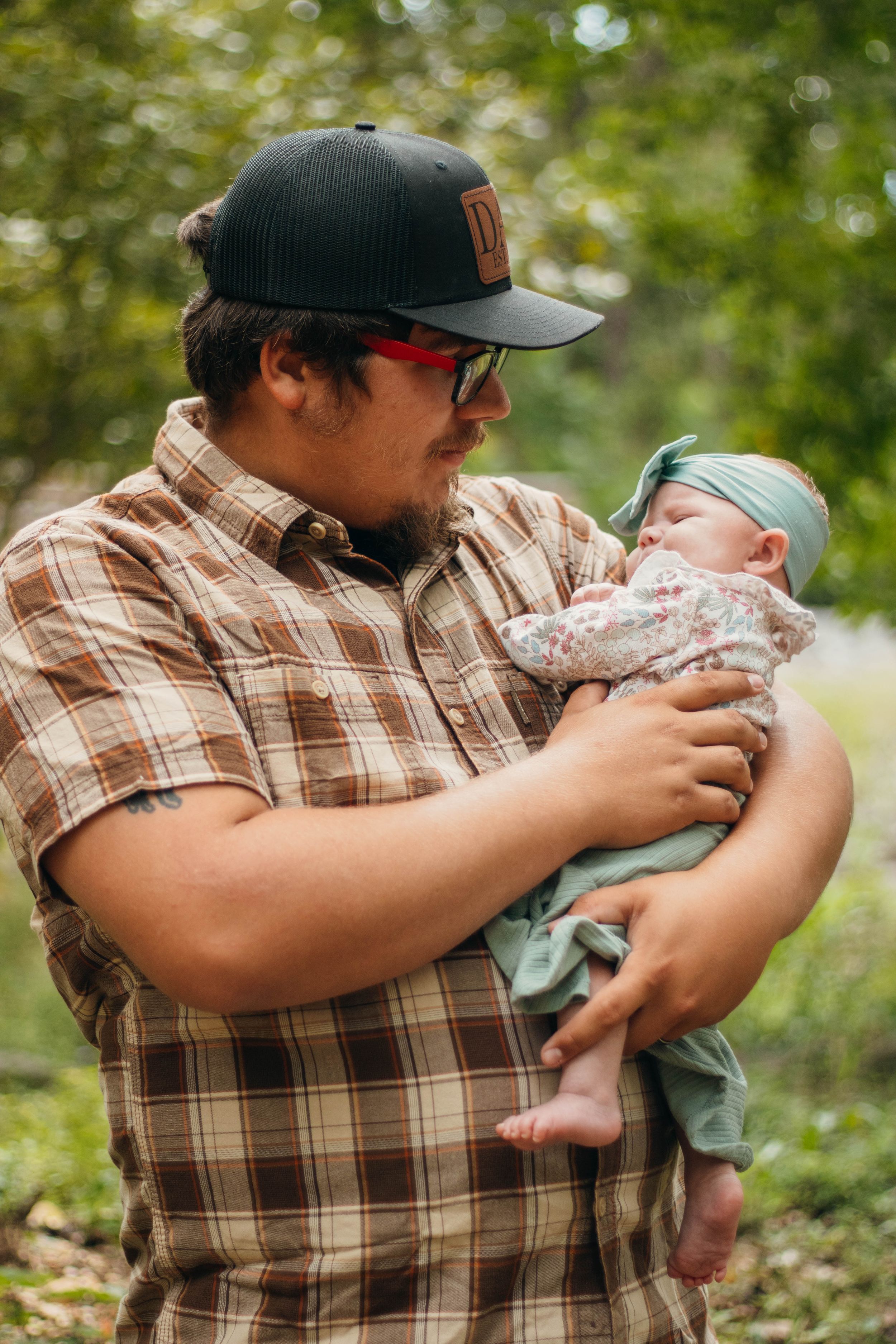 Person in plaid shirt and DAD cap tenderly holds a sleeping infant outdoors.