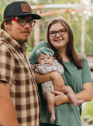 Happy family poses outdoors with their infant wearing a mint bow and floral romper.
