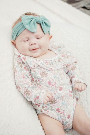 Smiling baby in floral onesie and mint bow headband laying on white blanket.