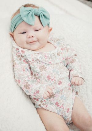 Happy baby wearing mint bow and floral outfit resting on white surface.