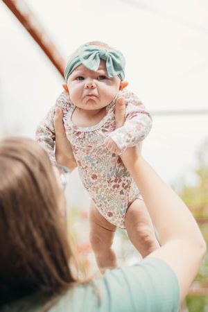 An infant wearing a mint green headband and floral onesie is held up in the air by unseen hands.