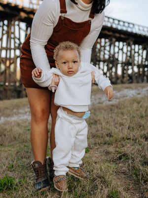 A toddler in white clothing takes early steps while holding an adult's hand near a rustic wooden bridge structure.