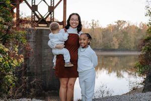 A couple in maroon and white outfits poses for autumn engagement photos near a lake at sunset.