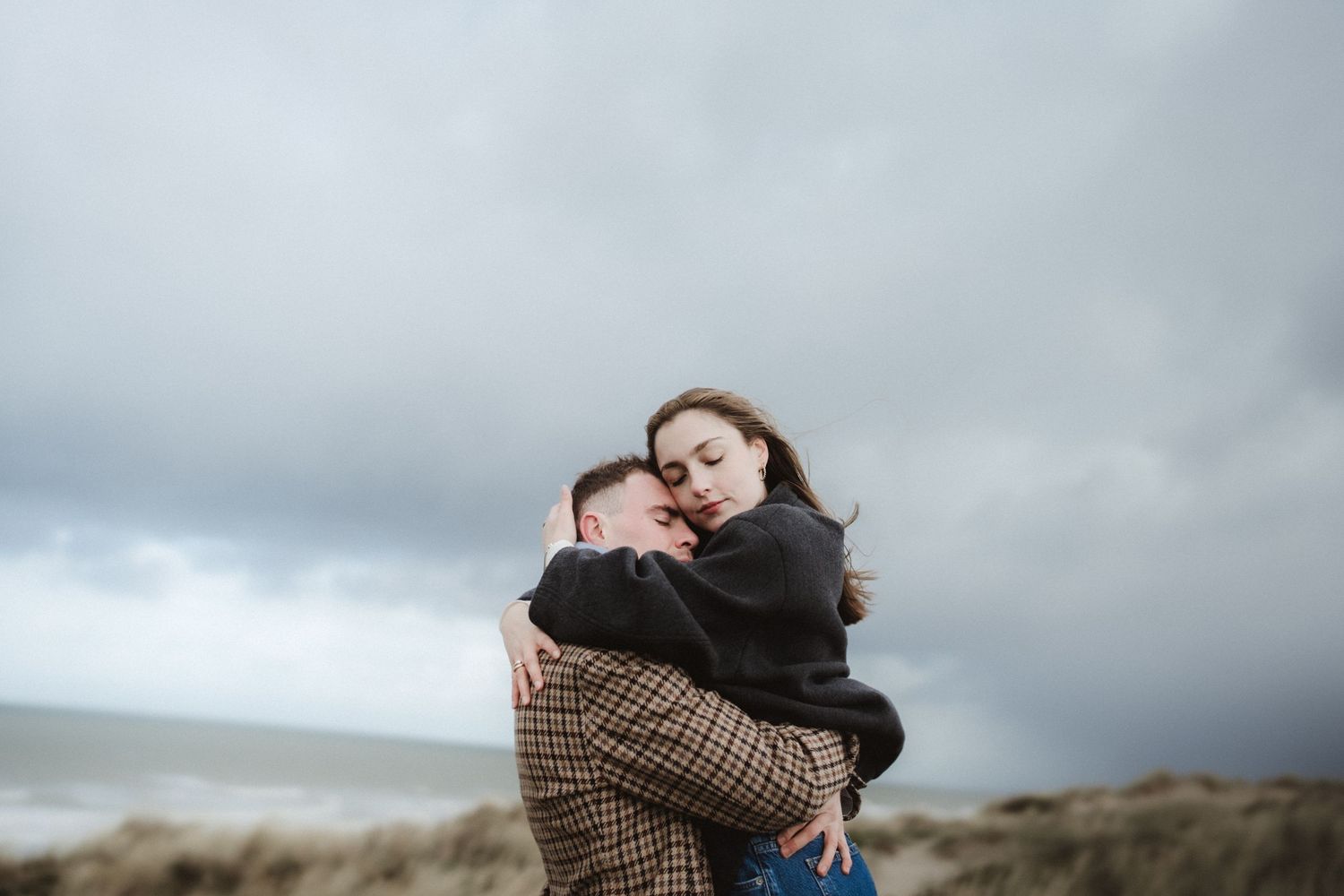 Couple sharing tender embrace on windswept coastal cliff under moody sky.