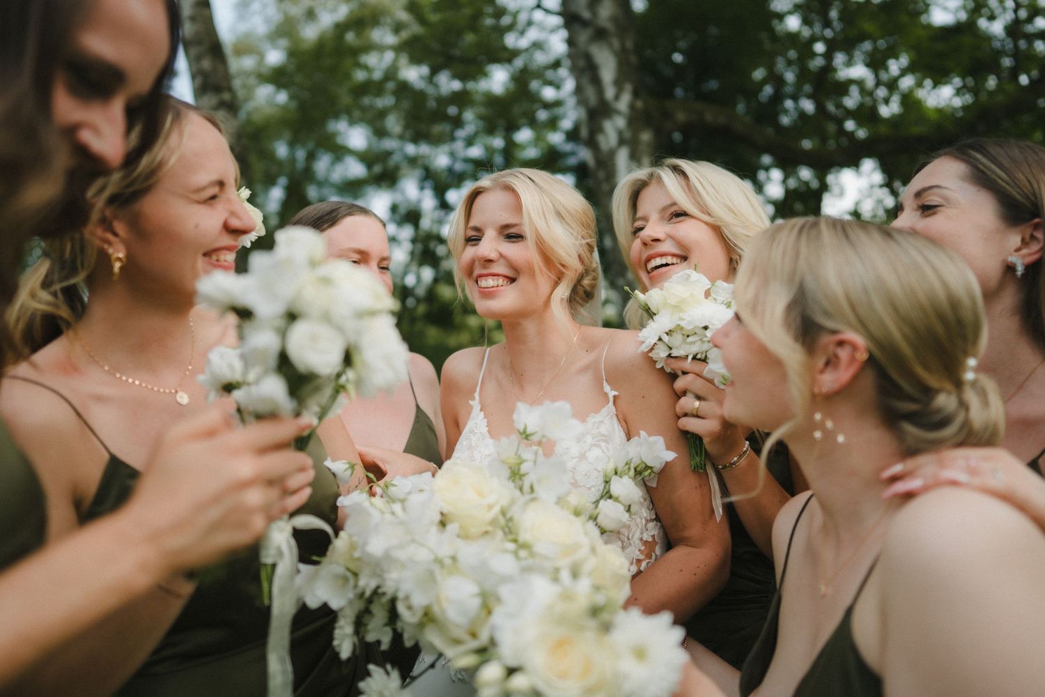 Group of wedding attendants holding bouquets share candid laughter on a sunny outdoor day.