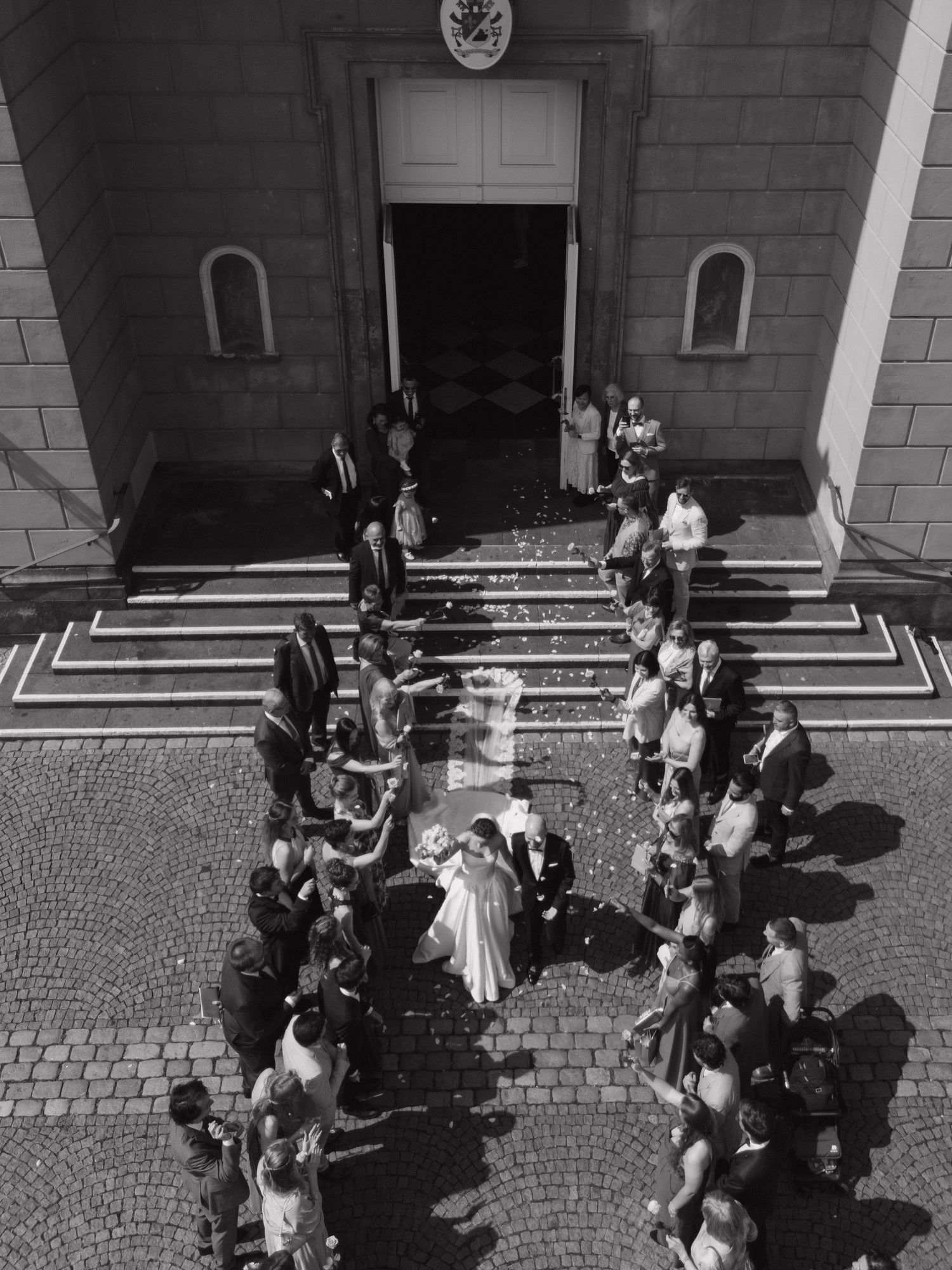 Aerial black and white photo of a wedding couple exiting a church, greeted by guests throwing confetti.