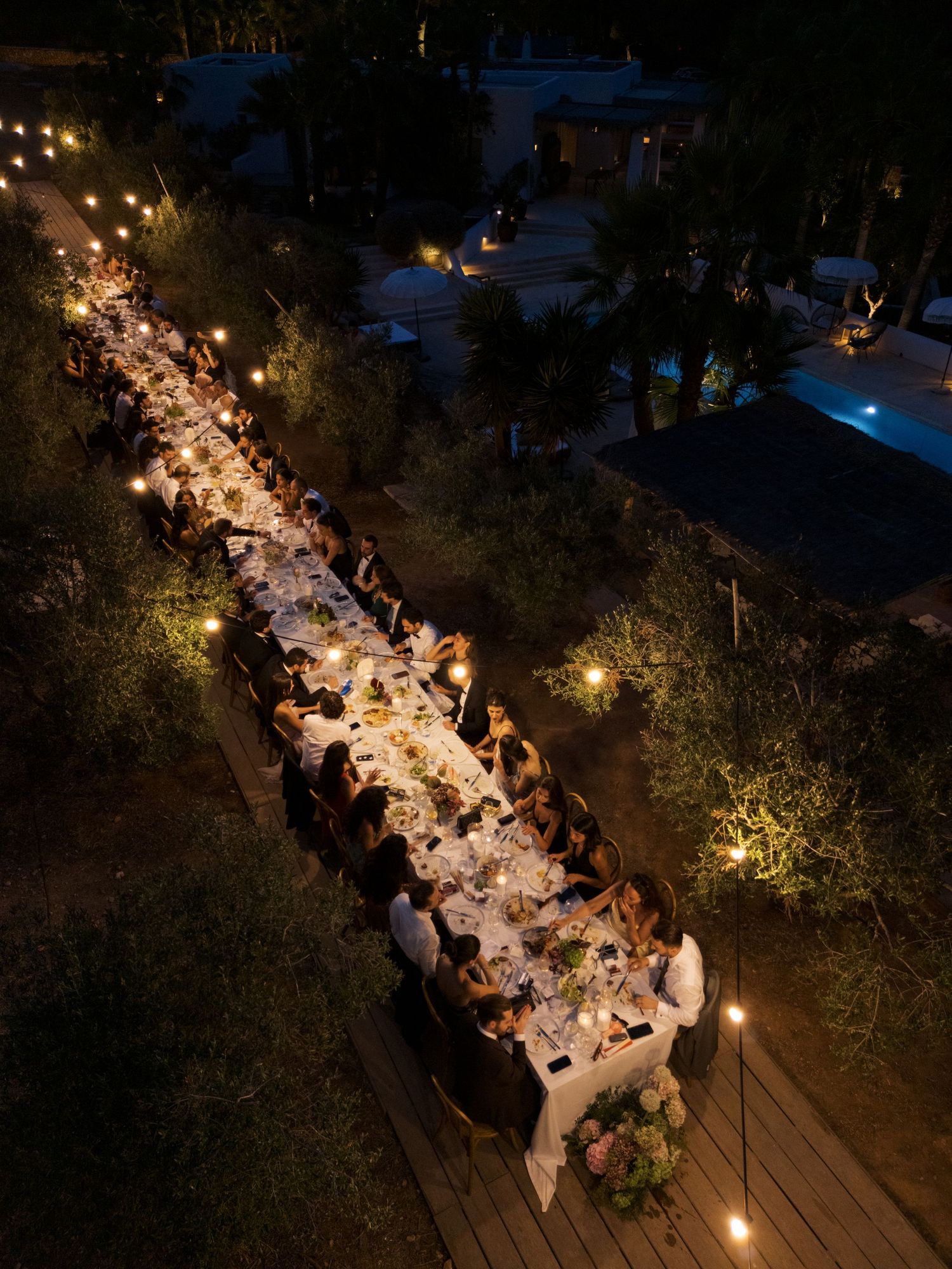 Aerial view of an elegant outdoor dinner party at night with long table, candlelight, and guests among trees.