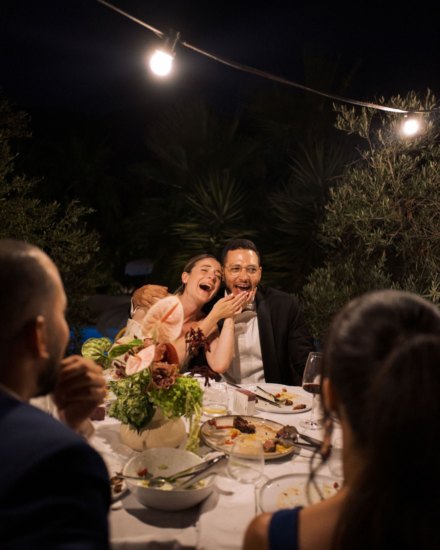 Couple laughing joyfully at outdoor evening wedding reception dinner table with string lights and floral centerpiece.