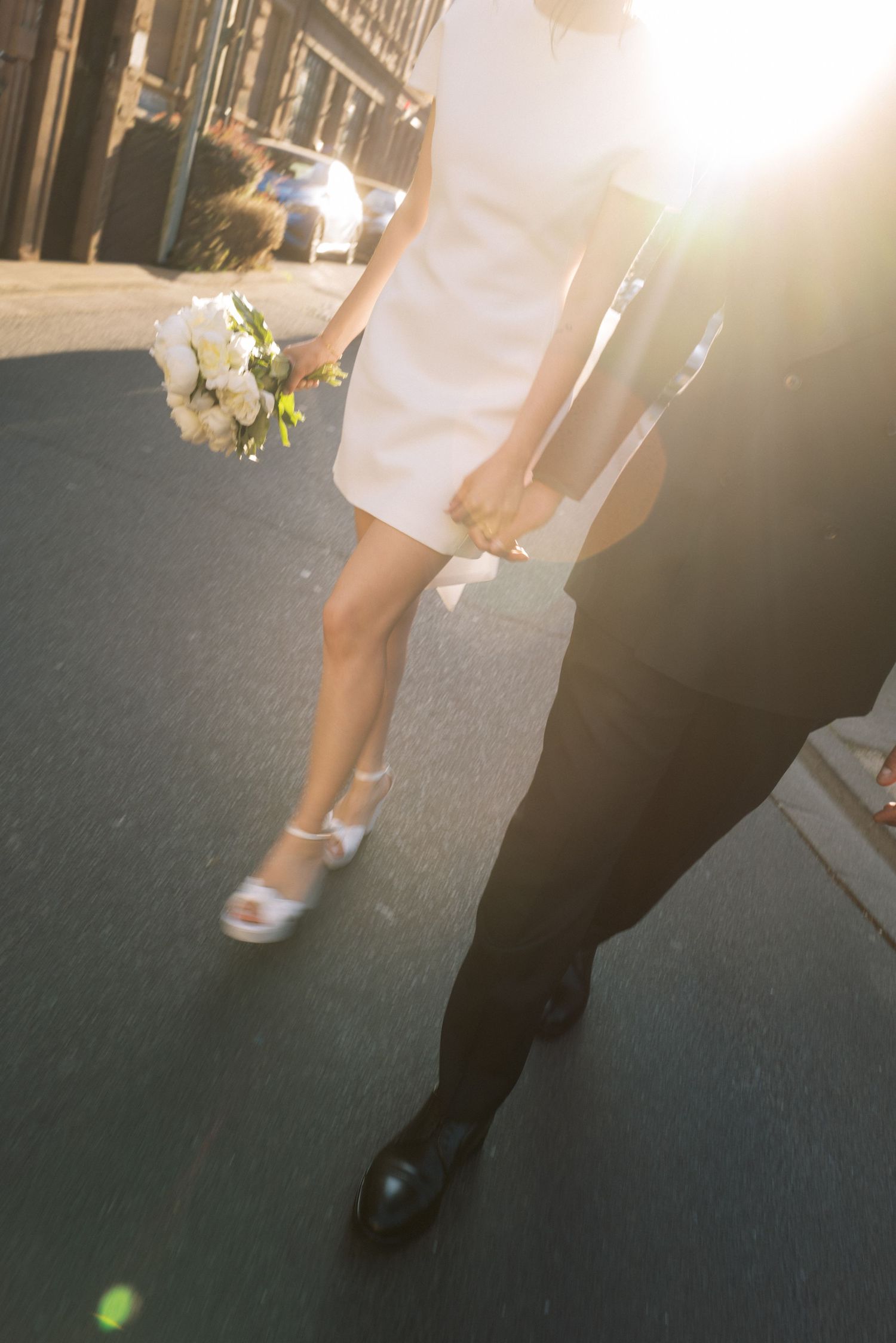 Bride in white mini dress holding bouquet walks with groom in black suit on sunny street.