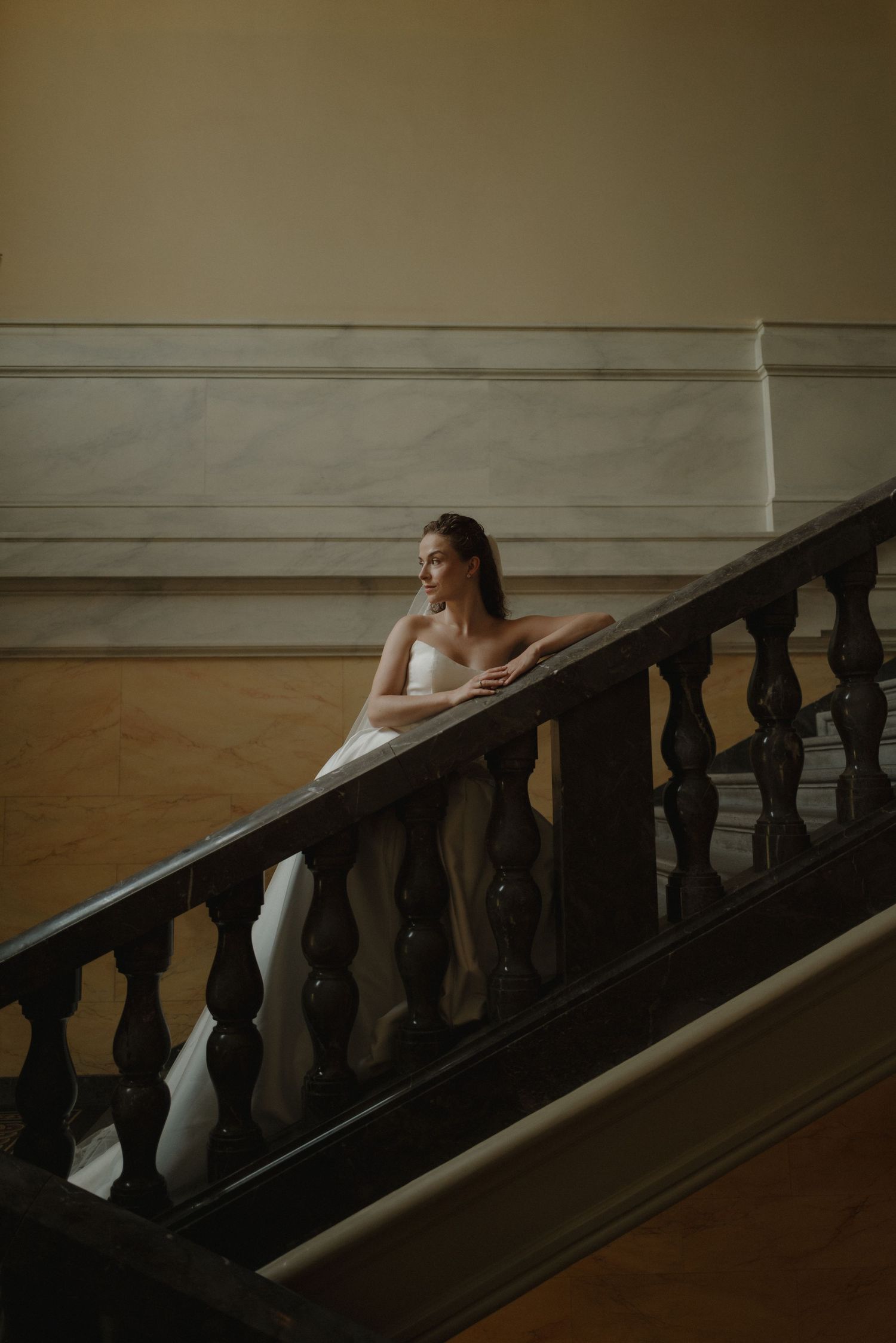 A bride in a white gown leans gracefully on a dark wooden staircase railing in an elegant classical interior.