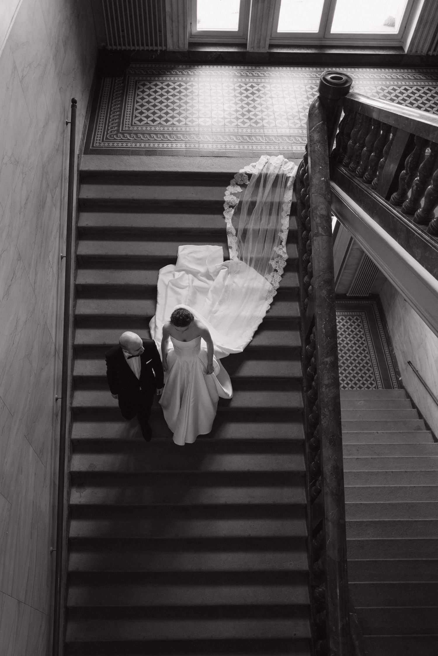 Aerial black and white photo of bride ascending grand staircase with escort, cathedral veil trailing elegantly.