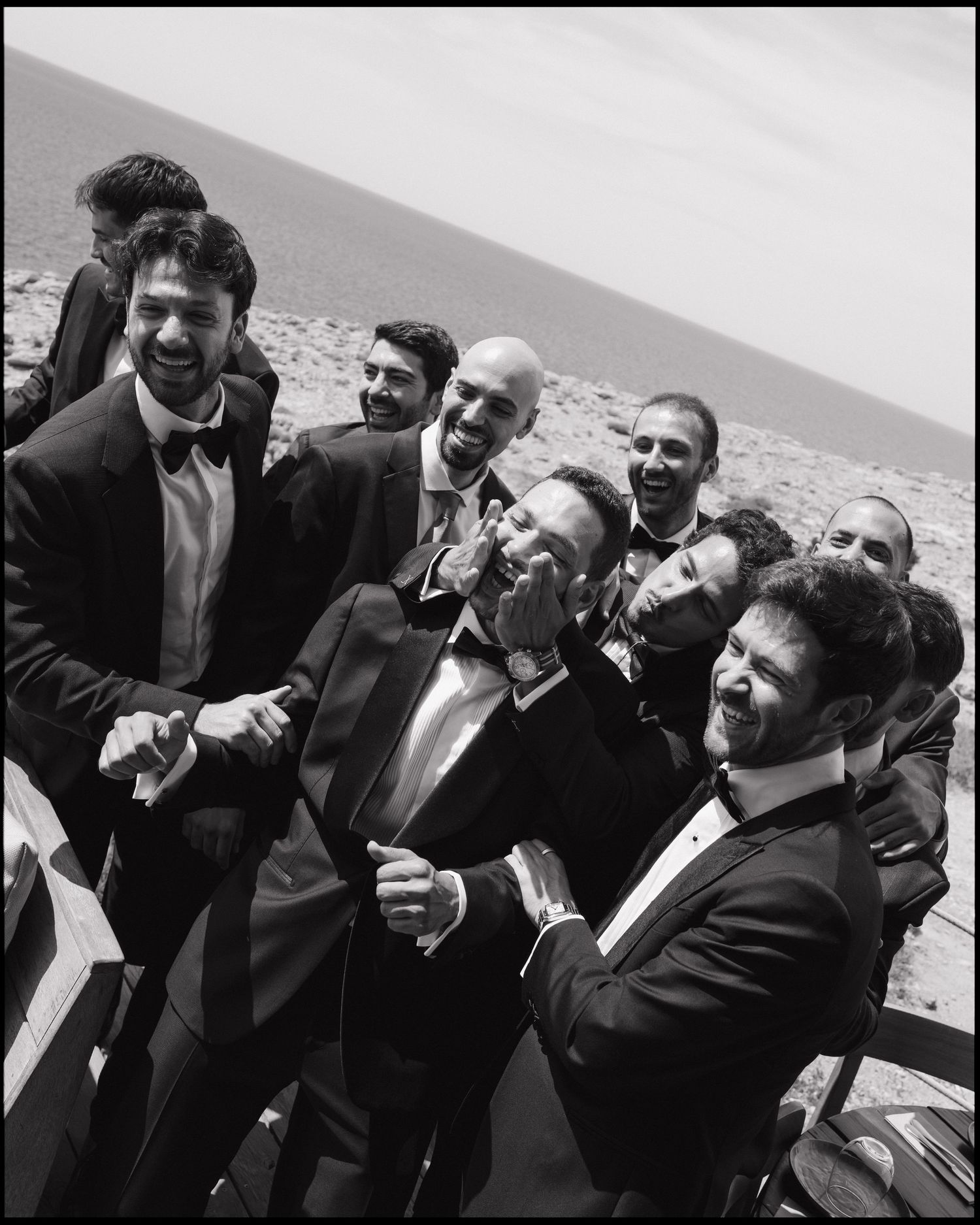 Black and white photo of groom laughing with groomsmen in tuxedos by the sea at a wedding celebration.