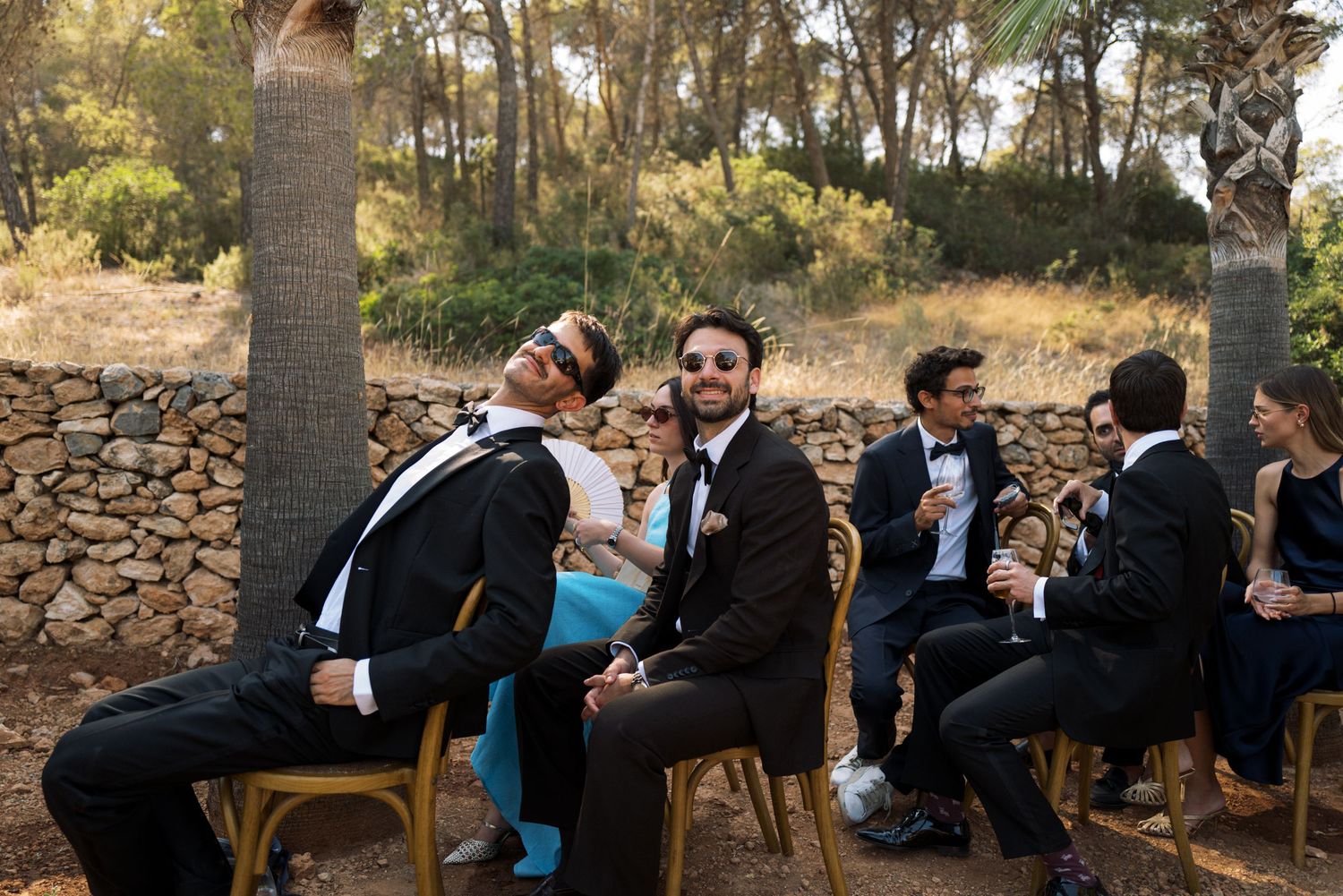 Wedding guests in formal attire laughing and relaxing on chairs outdoors near a stone wall and trees.