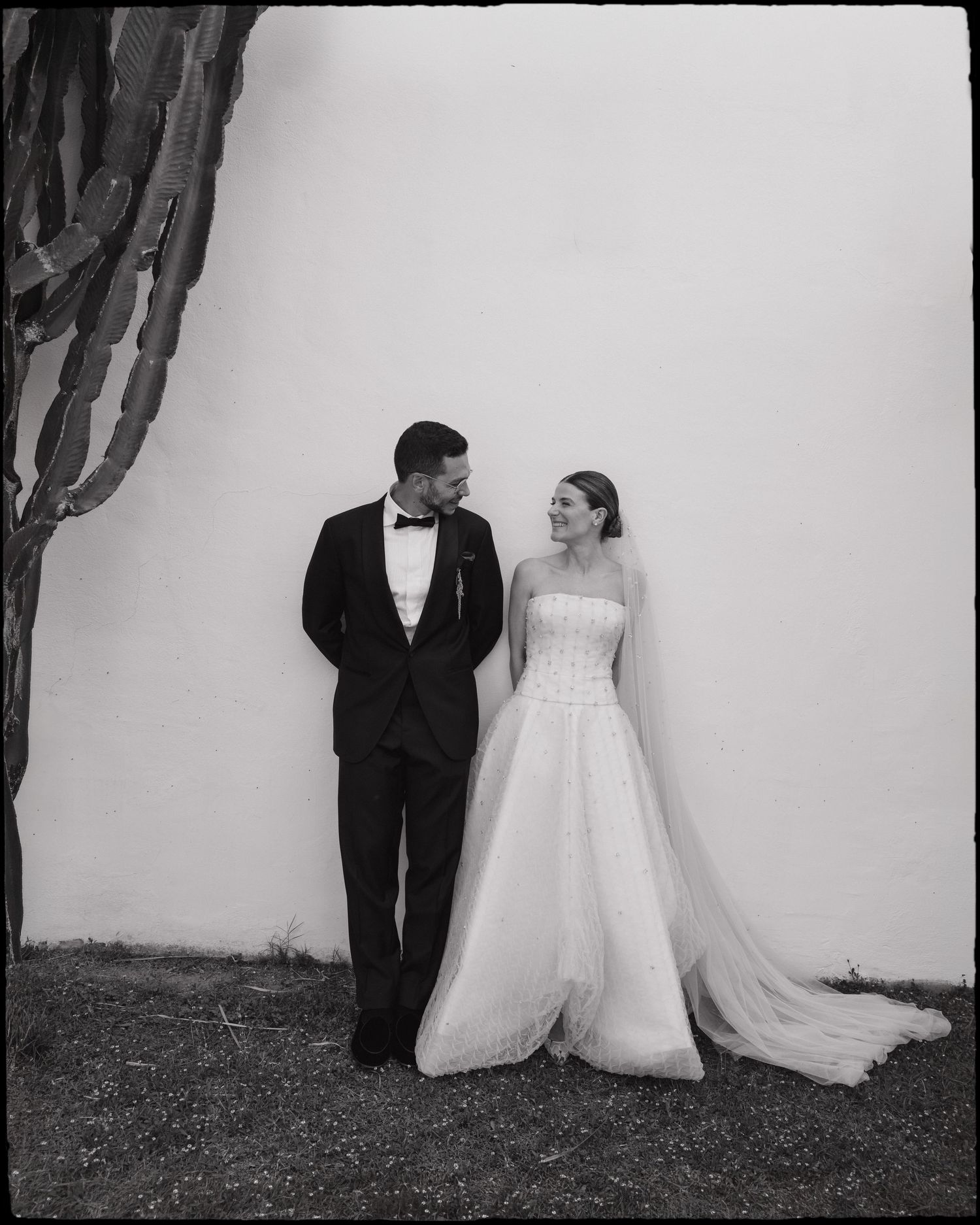 Black and white wedding photo of bride in strapless gown and groom in tuxedo standing by white wall with cactus.
