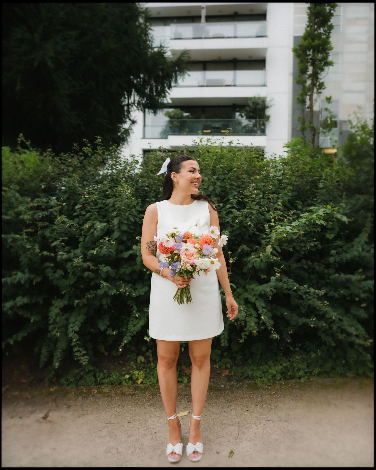 A person in a white dress holds a vibrant bouquet of flowers while standing in front of greenery.