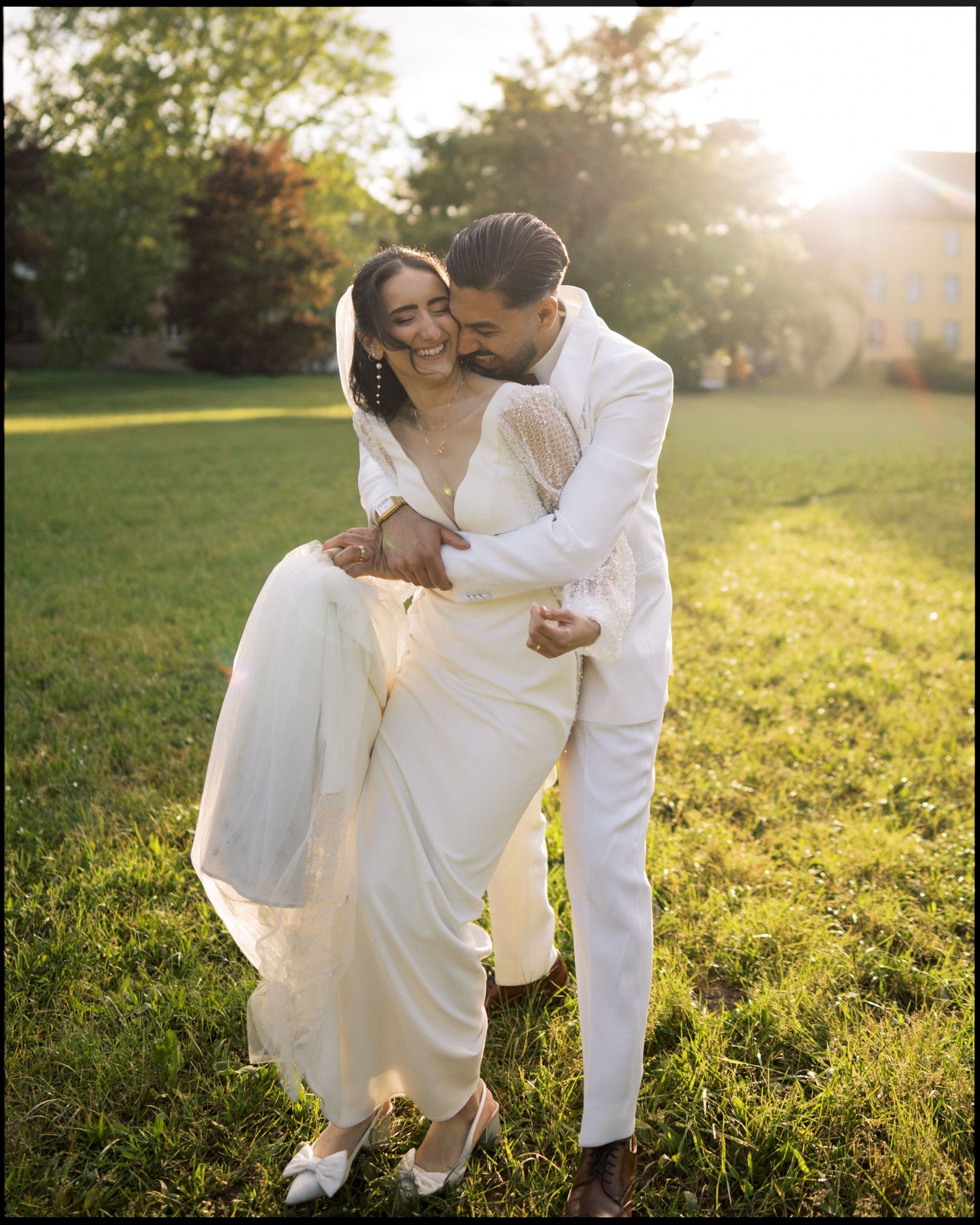 Couple in matching white outfits sharing a joyful embrace on green lawn at golden hour sunset.