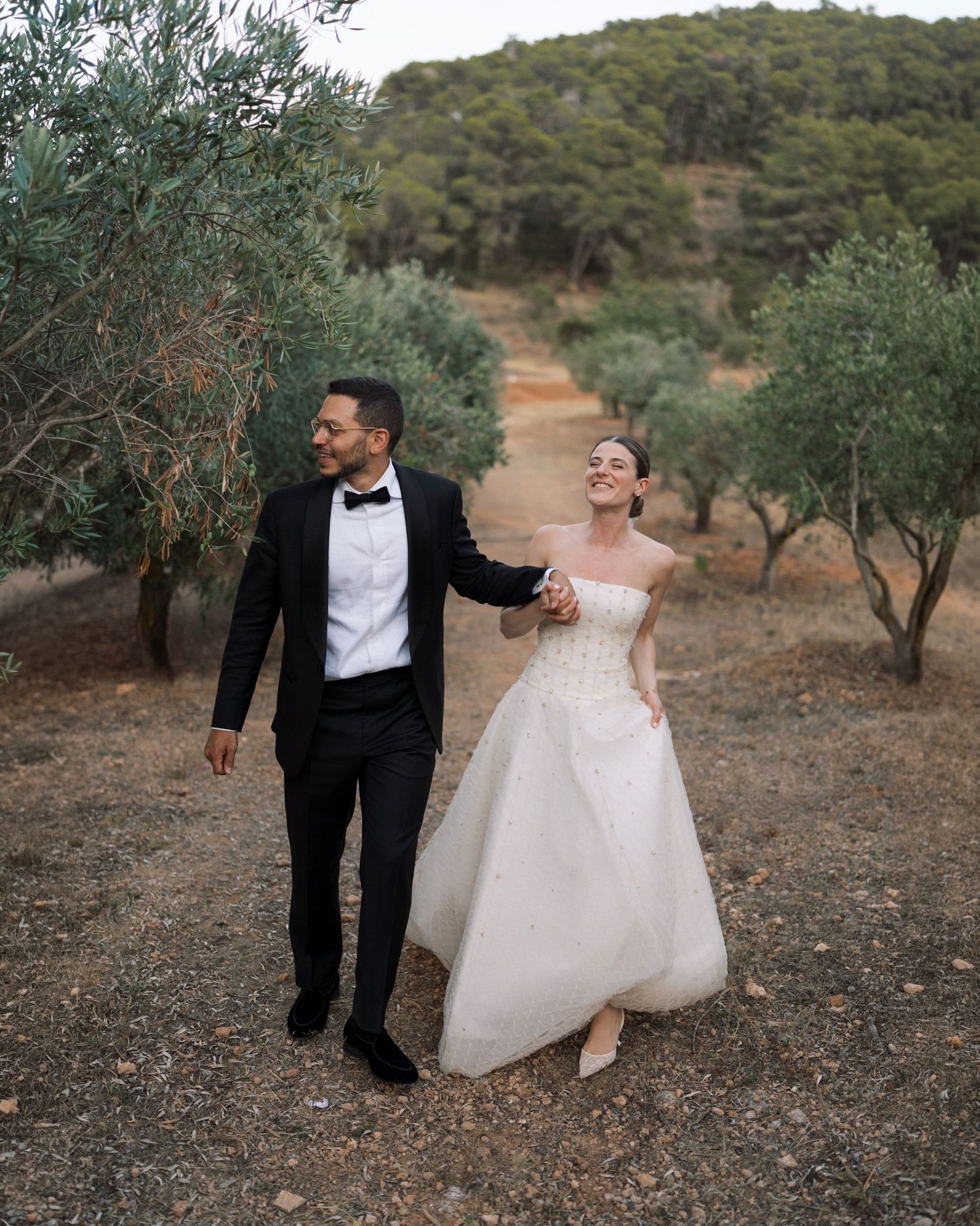 Bride and groom walking hand in hand through an olive grove, laughing joyfully on their wedding day.