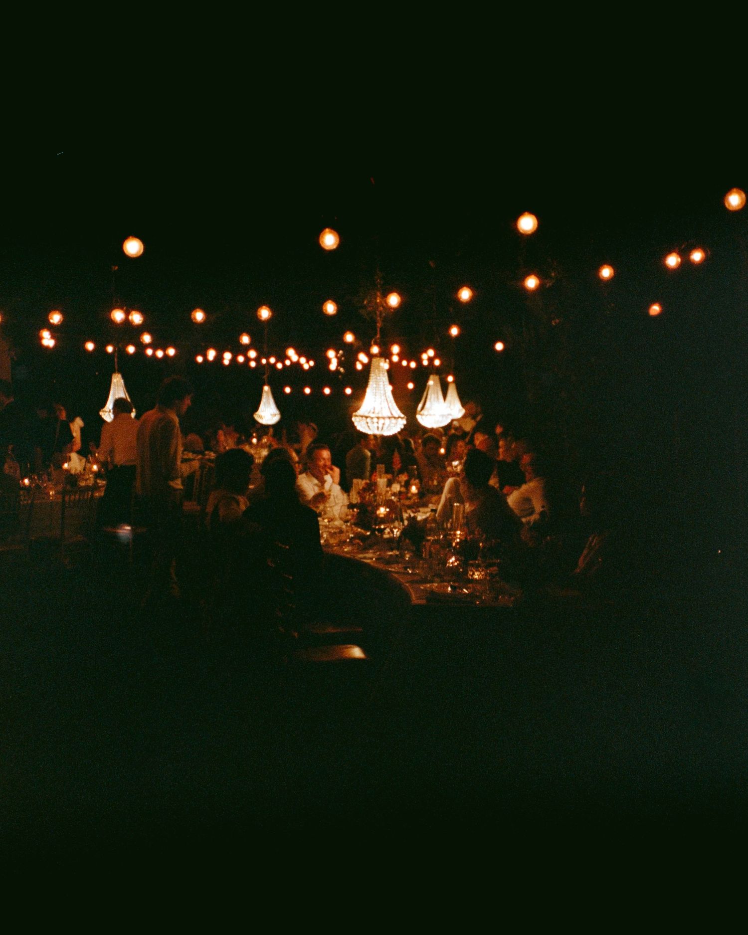 Romantic outdoor evening wedding reception with glowing chandeliers and string lights illuminating dining guests.
