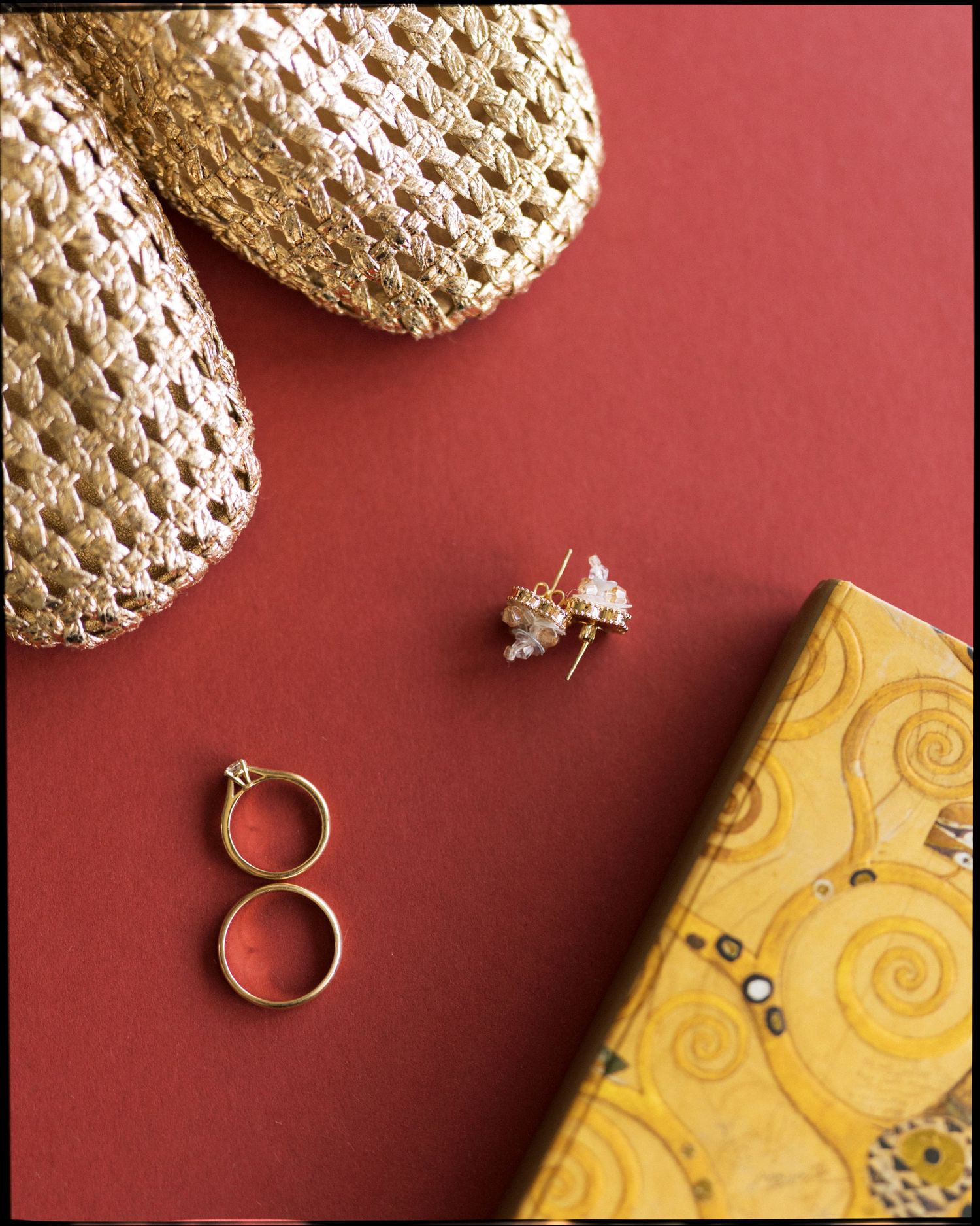 Gold woven slippers, crystal bee earrings, two gold rings, and a yellow Klimt notebook on a red background.