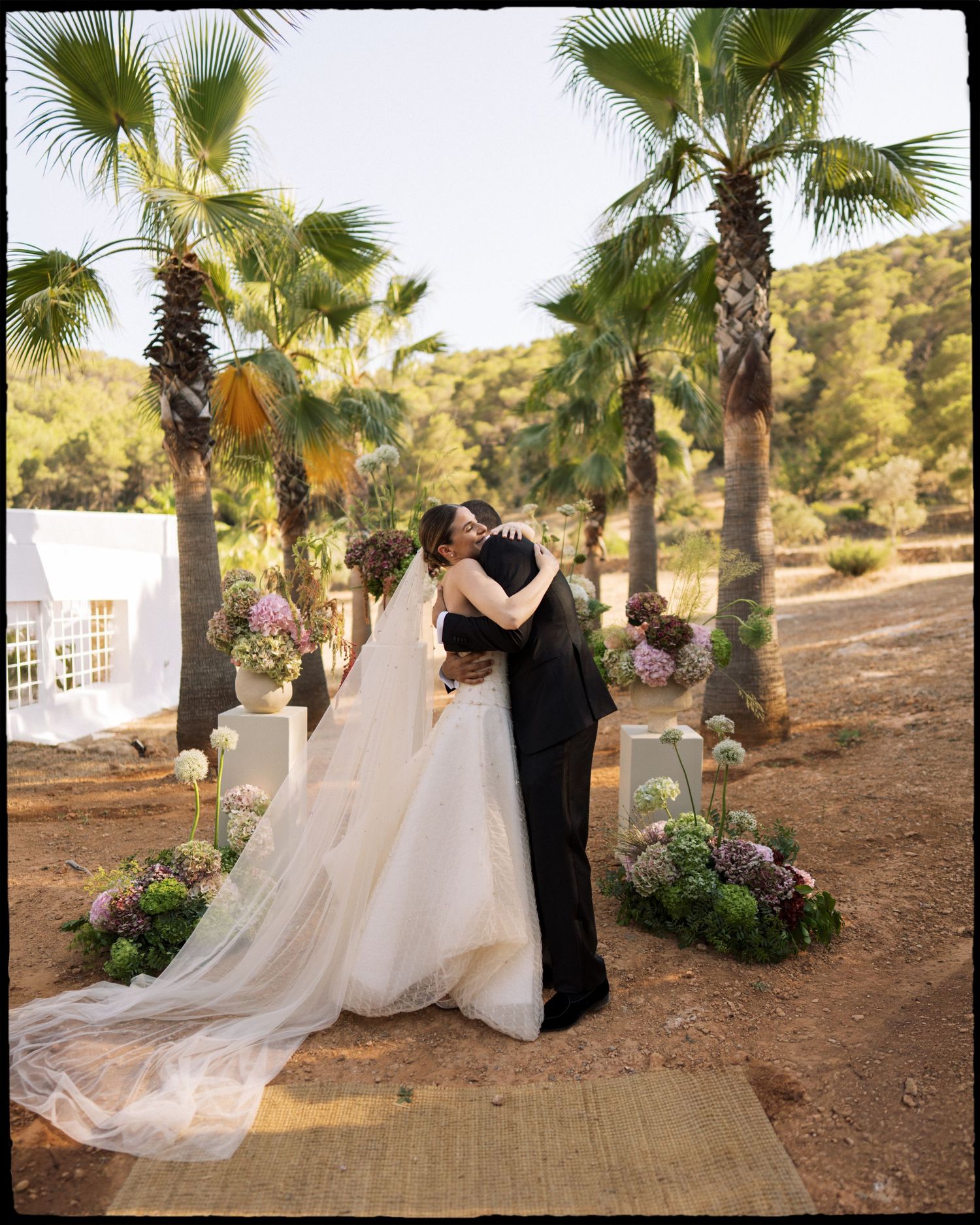 Bride and groom share first kiss at outdoor wedding ceremony surrounded by palm trees and floral arrangements.