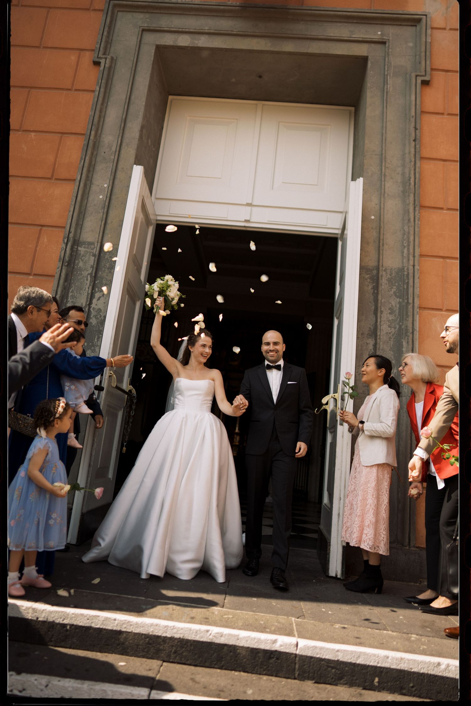 Bride and groom exit church doors as guests toss flower petals in joyful wedding celebration.