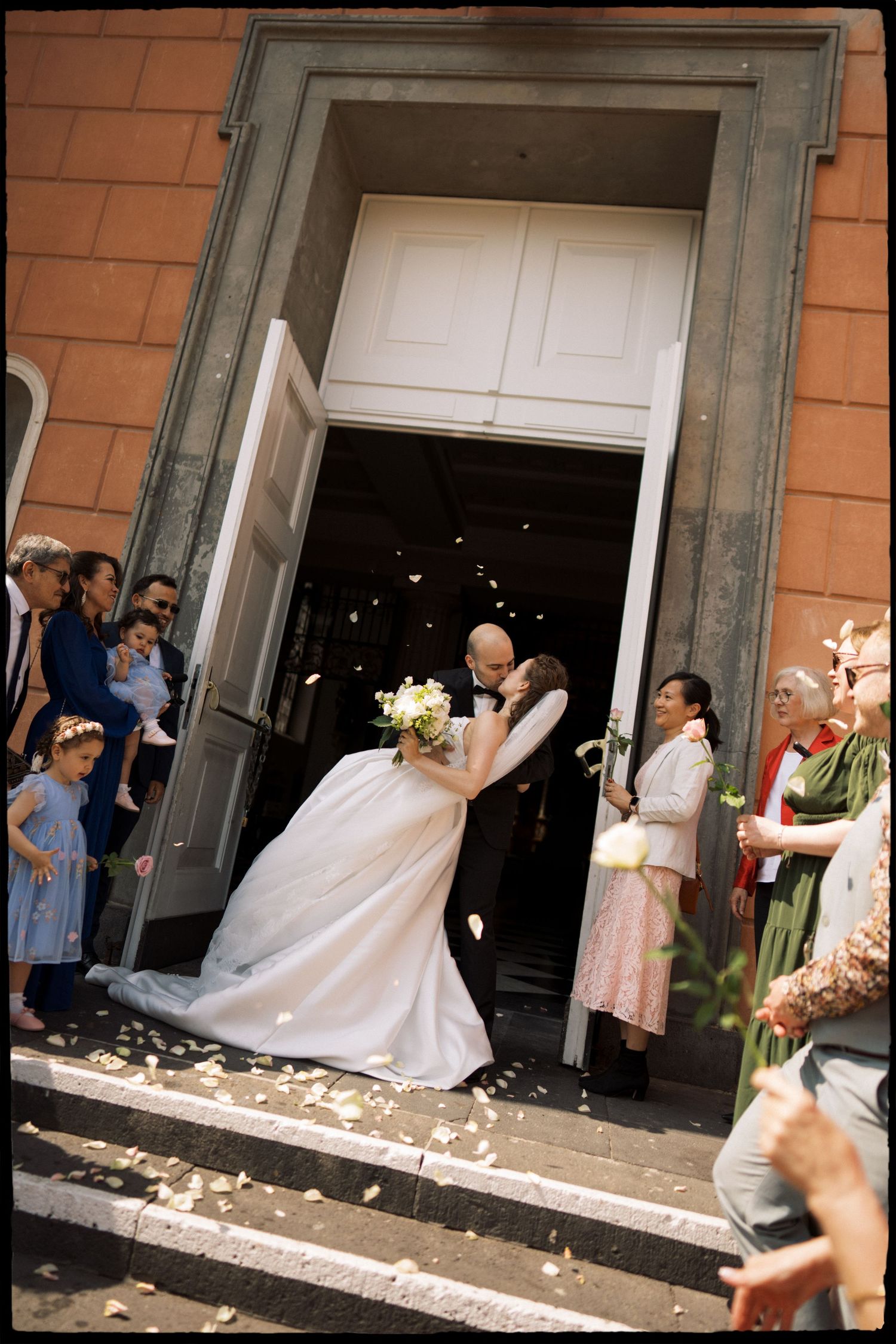 Newlyweds share a kiss at church entrance as guests celebrate with flower petals at outdoor wedding ceremony.