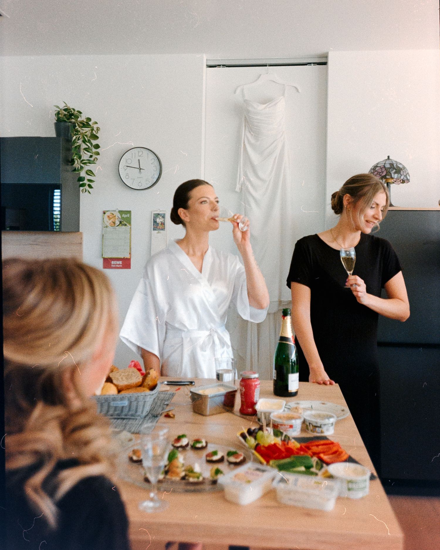 Bride in white robe with bridesmaids celebrating with champagne and food before wedding ceremony.