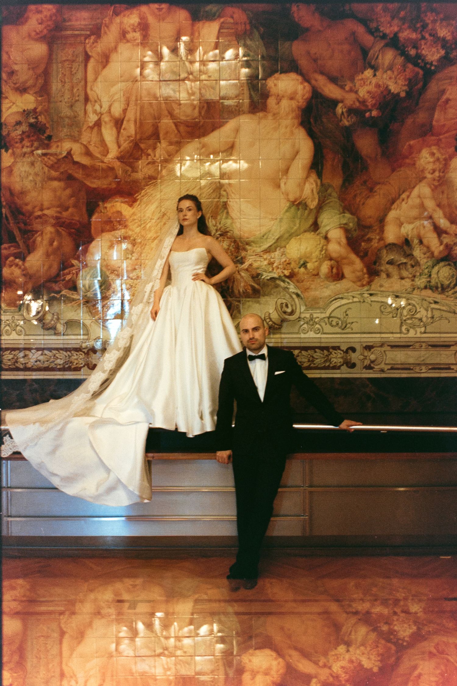 Bride in white gown stands above groom in tuxedo before ornate baroque mural in elegant venue.