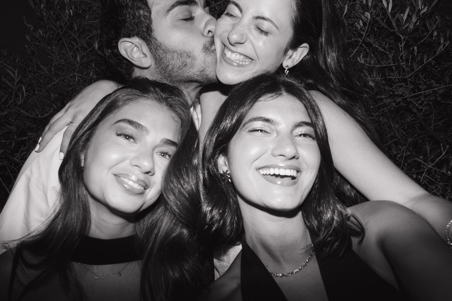 Black and white photo of three smiling young women and a man kissing one on the cheek outdoors at night.