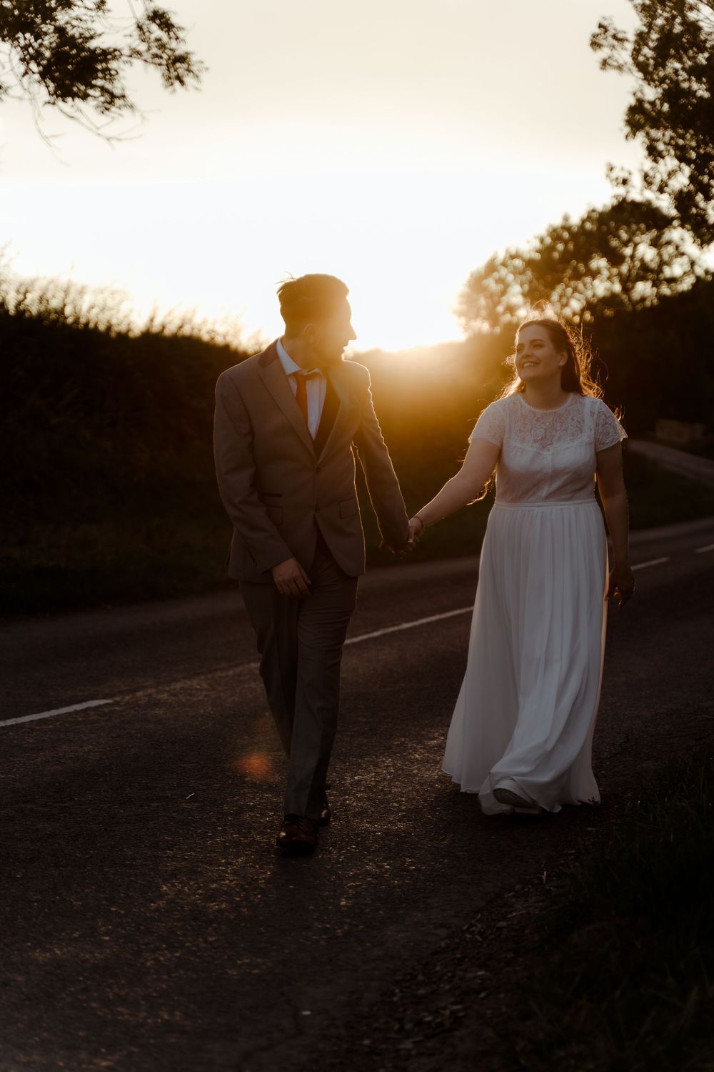 A romantic sunset silhouette of a couple walking together on a path, the bride in white and groom in a suit.