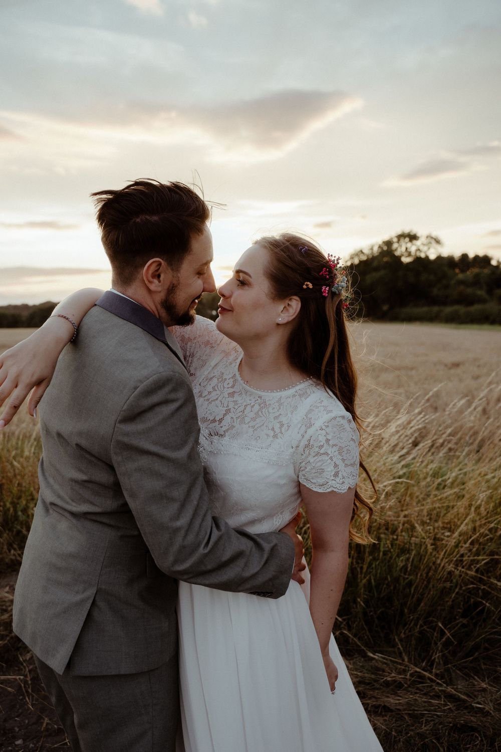 Romantic couple embraces in a golden wheat field at sunset wearing wedding attire.