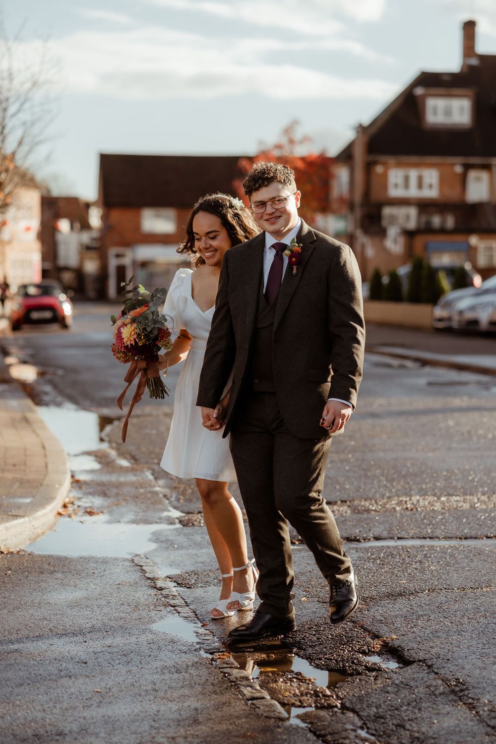 A couple in wedding attire walks together on a paved street while holding autumn-colored floral bouquets.