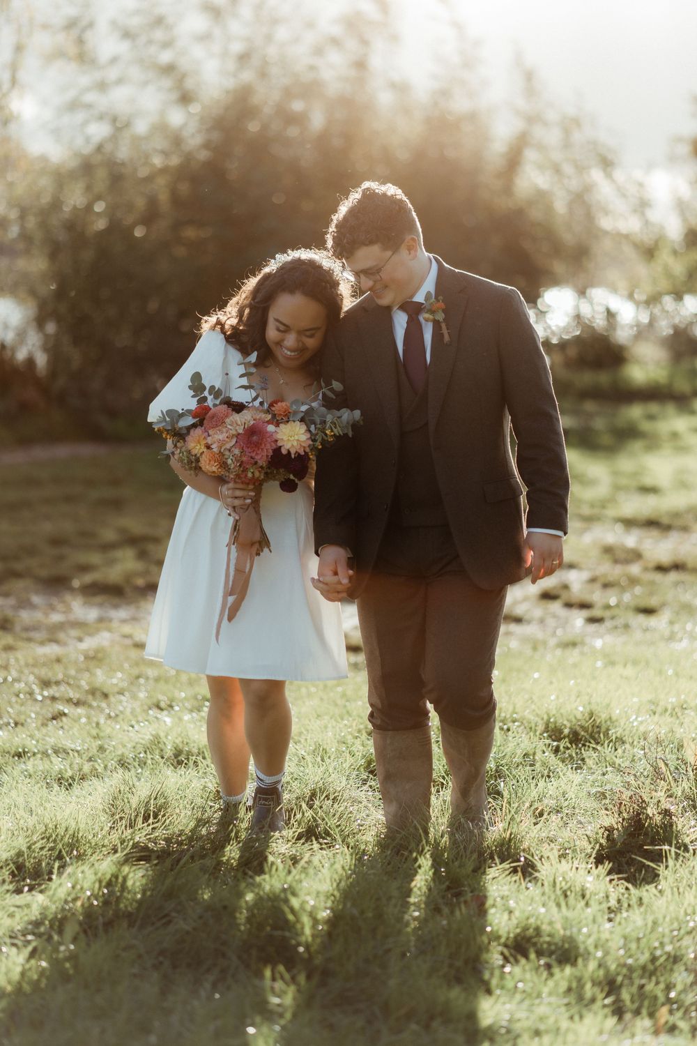 A couple walks together through a sunlit meadow during golden hour holding colorful flower bouquets.