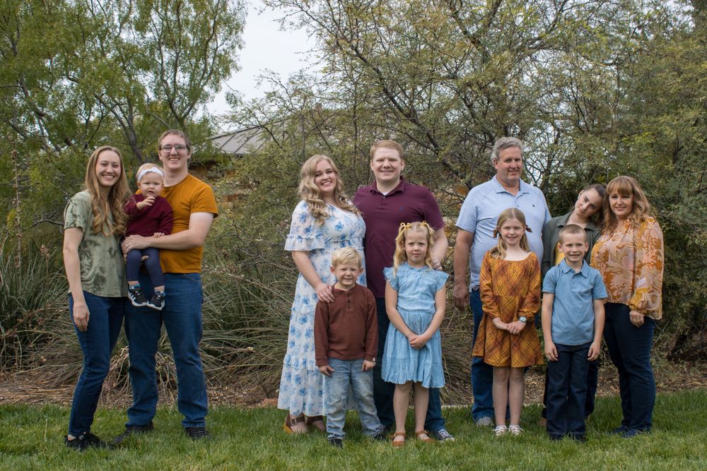 A large extended family of ten people poses together outdoors on a green lawn with trees behind them.
