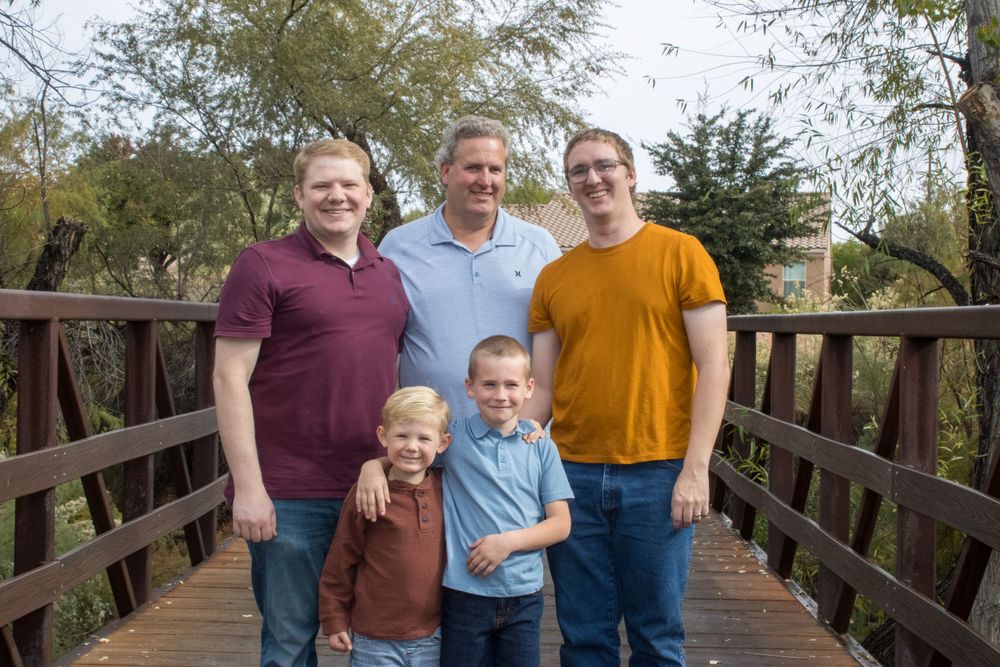 Three men and two young boys smile together on a wooden bridge in a family portrait.