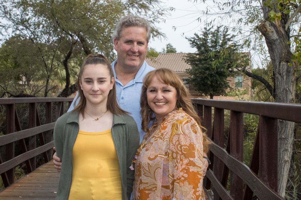 A teenage girl poses with her parents on a wooden bridge in an outdoor family portrait.