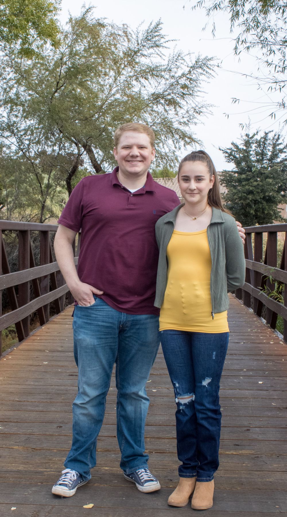 A young man in maroon polo and a woman in yellow top stand together on a wooden bridge outdoors.