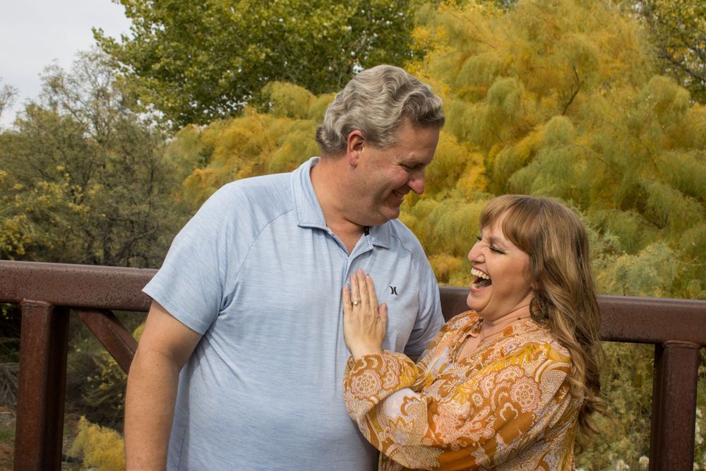 A cheerful middle-aged couple laughs together on a wooden deck surrounded by colorful autumn trees.