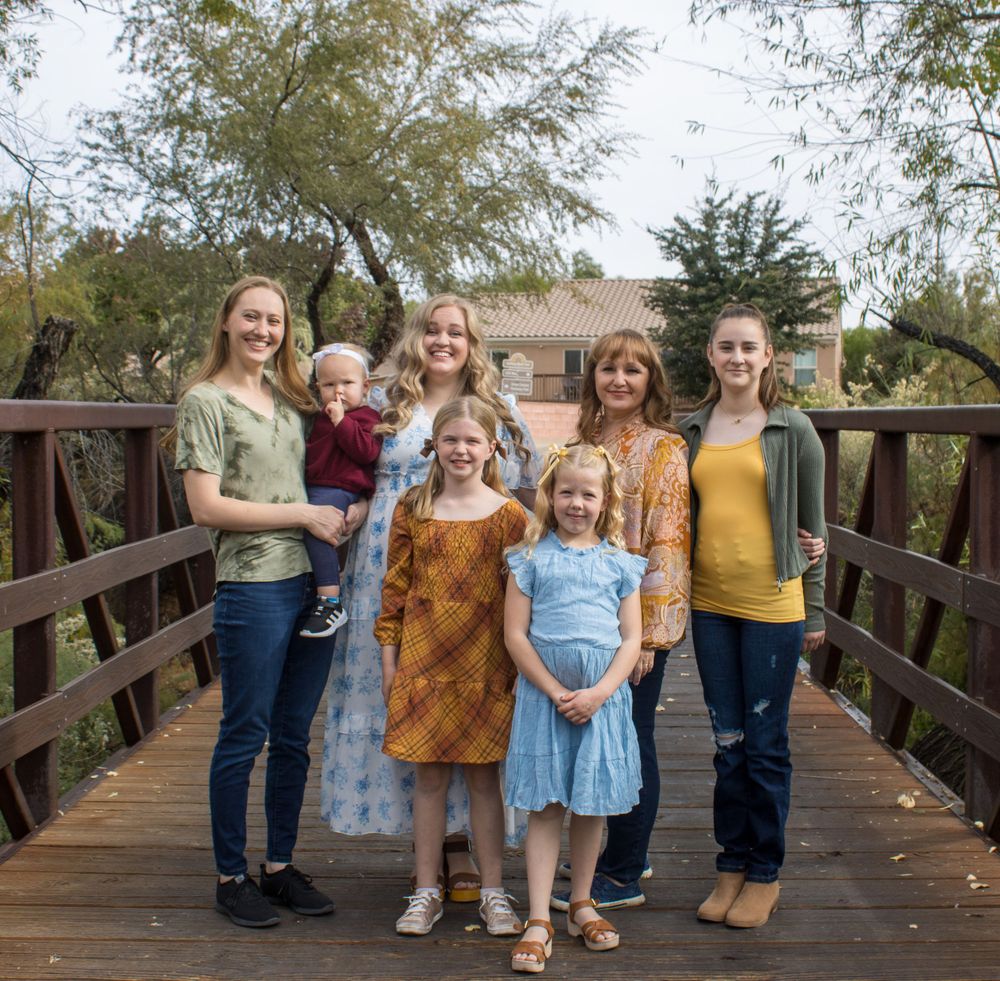 Group of seven women and girls smiling together on a wooden bridge outdoors.