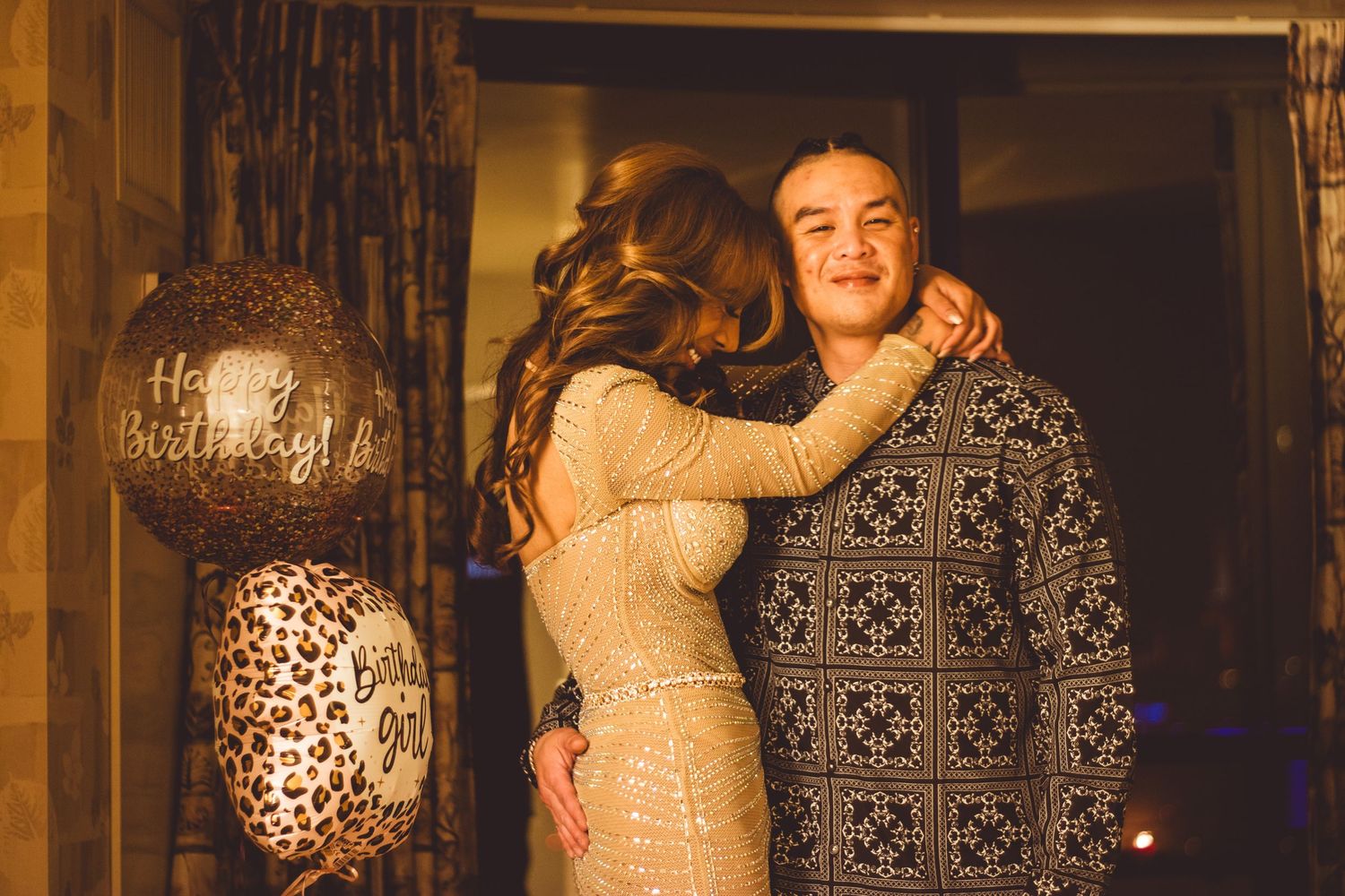 A couple embraces near Happy Birthday balloons in a warmly lit celebratory setting.