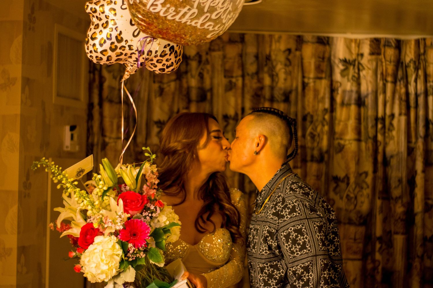 A couple shares a romantic kiss surrounded by birthday balloons and a colorful floral bouquet.