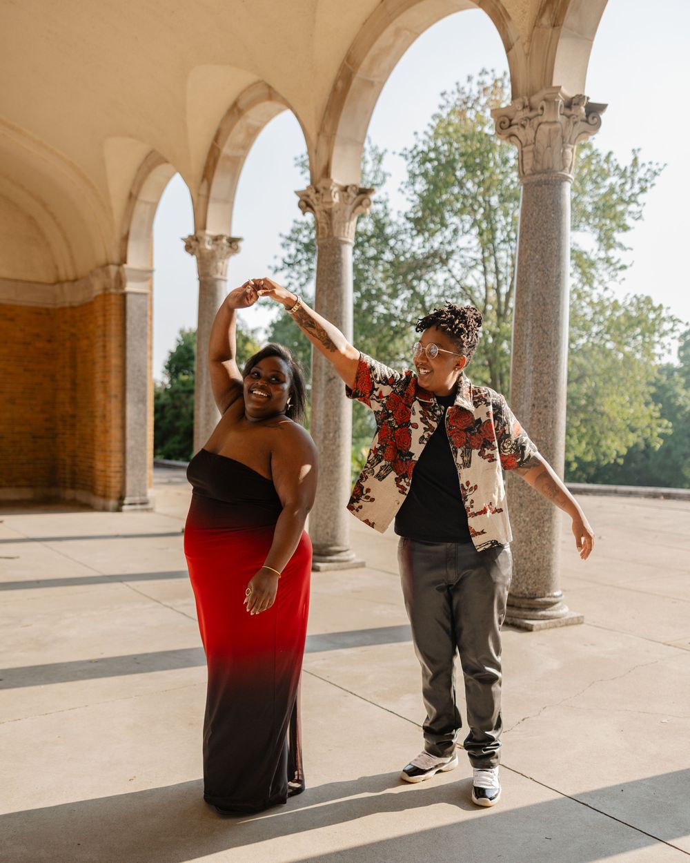 Couple dancing joyfully under stone arches, woman in red ombre gown, man in floral shirt, outdoor venue.