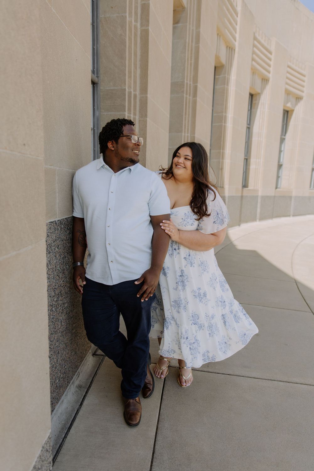 A couple poses against a stone wall outdoors, woman in floral dress and man in white shirt smiling together.