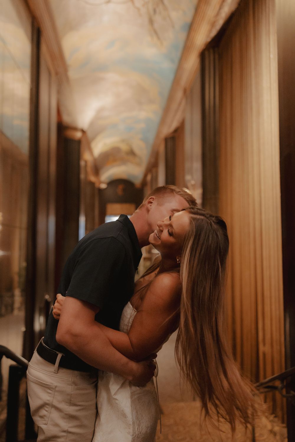 A couple shares a romantic embrace and kiss in an ornate hallway with painted ceilings and golden curtains.
