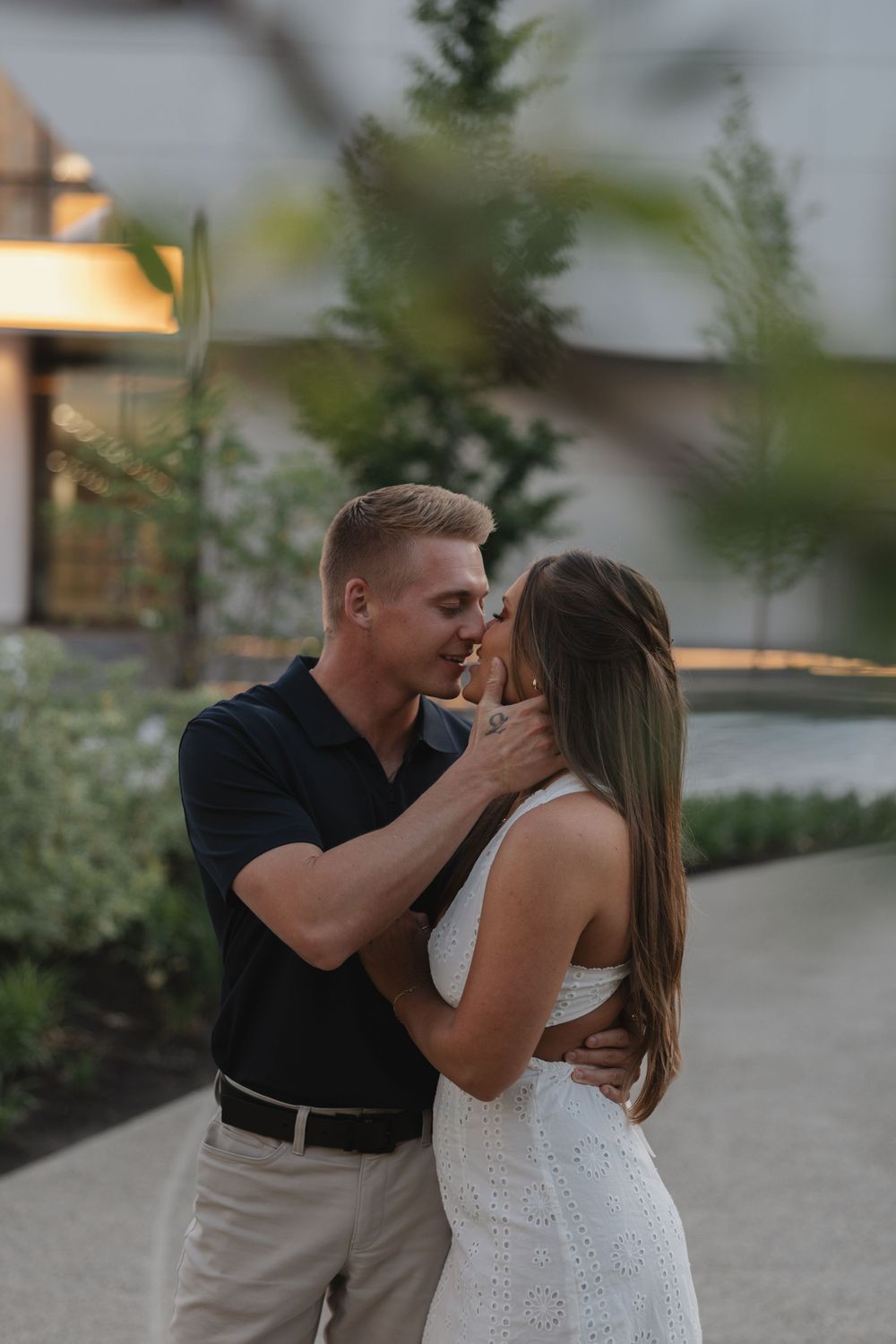 A couple shares a romantic moment outdoors at dusk, man in navy polo gently holding womans face in white lace dress.