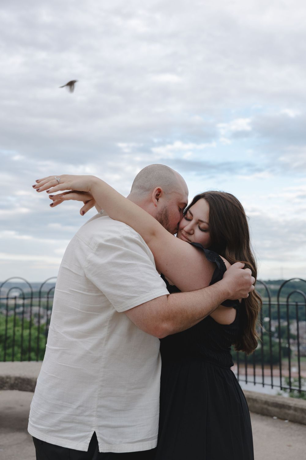 A couple embraces tenderly on an outdoor overlook, with a cloudy sky and green landscape in the background.